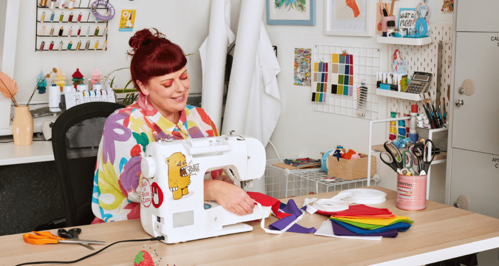 A smiling redhead is expertly sewing a colorful fabric with a sewing machine in a vibrant craft room. Spools of thread, scissors, and a pincushion are neatly arranged on the table. Art supplies are meticulously organized in the background, highlighting a passion for creative sewing projects.