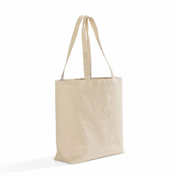 A plain beige canvas tote bag with long handles is standing upright against a white background. The bag is unadorned and has a simple, minimalist design.