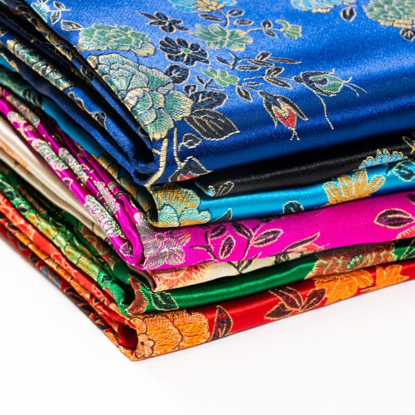 A stack of colorful, folded brocade fabrics with intricate floral patterns. The fabrics include shades of blue, pink, red, and orange, and display detailed gold, green, and black embroidery. The fabrics are neatly arranged on a white background.