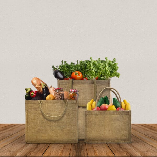 Three burlap bags filled with various fruits, vegetables, and jars are placed on a wooden surface against a plain light background. The bags contain items like lettuce, tomatoes, eggplants, bread, bell peppers, and bananas.