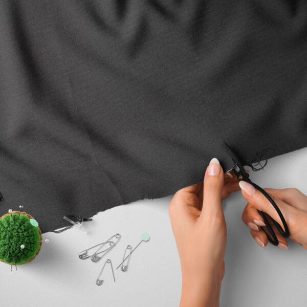 Hands cutting a piece of dark gray fabric with black scissors. There is a small pincushion with green pins and several safety pins scattered on the white surface nearby.
