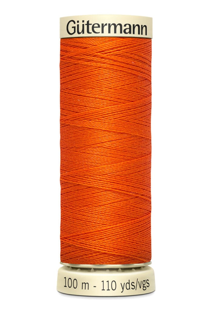 A spool of Gütermann orange sewing thread is shown, labeled with "100 m - 110 yds/vgs." The spool is off-white with the brand name printed at the top. The thread is tightly wound around the spool.
