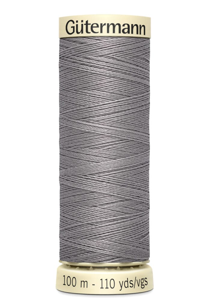 Spool of Gütermann thread, gray in color, with labeling indicating 100 meters or 110 yards of thread. The spool is upright, with beige ends and crisscrossing thread visible.