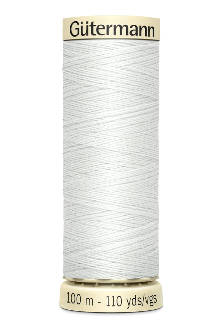 A spool of Gütermann sewing thread sits vertically. It is off-white and has a label indicating 100 meters or 110 yards of thread. The spool is cylindrical with a beige cap.