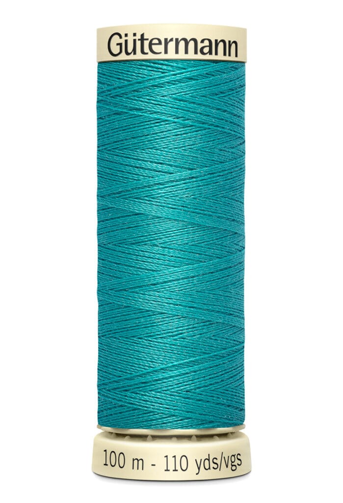 A spool of Gütermann sewing thread in teal is standing upright. The label indicates 100 meters, 110 yards. The spool is cream-colored with black text.