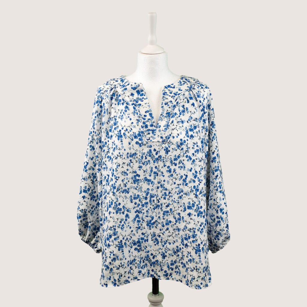 White blouse with a blue floral pattern displayed on a mannequin. The long-sleeve blouse features a split neckline and a loose, flowing fit, set against a plain light grey background.