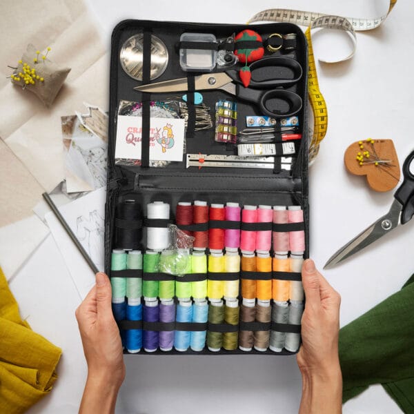 Hands holding an open sewing kit containing colorful spools of thread, scissors, measuring tape, fabric pieces, and various sewing tools. A variety of fabric swatches and pins are visible in the background.