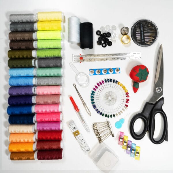 A sewing kit displayed on a white background, featuring various spools of thread in different colors, needles, measuring tape, buttons, safety pins, a seam ripper, scissors, and a red tomato pin cushion.