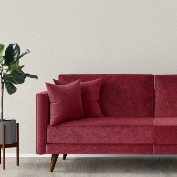 A modern living room features a red velvet sofa with two matching pillows, set against a textured beige wall. Beside the sofa is a tall potted plant with glossy green leaves, placed in a sleek grey planter on light wood flooring.