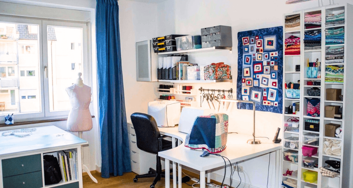 A bright sewing room with a mannequin near a window, two desks with sewing machines, shelves with neatly organized fabrics and sewing supplies, and a colorful quilt hanging on the wall. The room has white walls and wooden flooring.