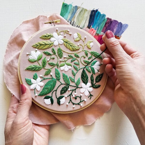 A person holds an embroidery hoop displaying an intricate floral design with green leaves and white flowers. Spools of colorful embroidery thread are visible in the background.