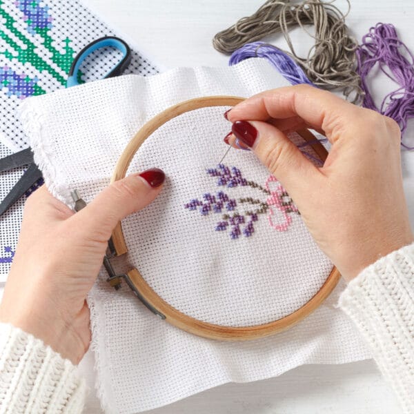 Hands embroidering a lavender and pink floral design on white fabric using a wooden hoop. Various skeins of thread, scissors, and a pattern are visible on the table.