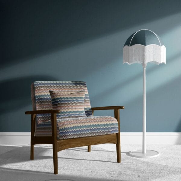 A wooden armchair with a zigzag patterned cushion is placed beside a tall, white floor lamp with a fringed, multicolored shade. The background features a solid teal wall and a light gray carpeted floor.