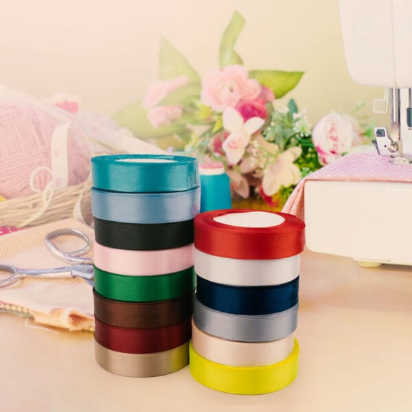 Stacked colorful ribbons are prominently displayed on a table. In the background, there are pink flowers, scissors, a spool of pink yarn, and part of a sewing machine. The scene conveys a crafting or sewing project environment.