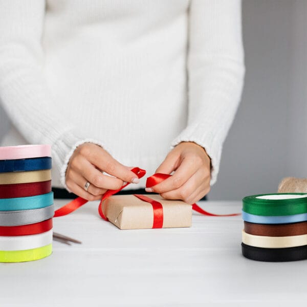 A person in a white sweater wraps a gift with red ribbon on a white table. Various colored ribbon rolls are stacked on the left. Scissors are visible next to the ribbons.