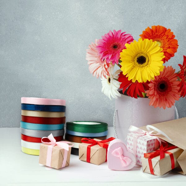 A white bucket holds vibrant gerbera daisies in pink, orange, red, and yellow. Nearby, colorful rolls of ribbon, small gift boxes tied with bows, and a pink heart-shaped box are arranged on a white surface against a gray background.