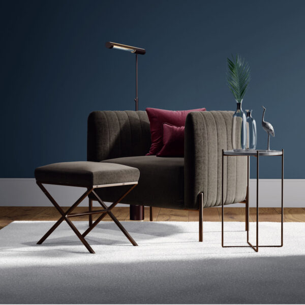 A modern living room with a dark green velvet armchair, matching ottoman, side table with glass vases and a bird sculpture, a floor lamp, and a blue wall background. A light rug covers a wooden floor.