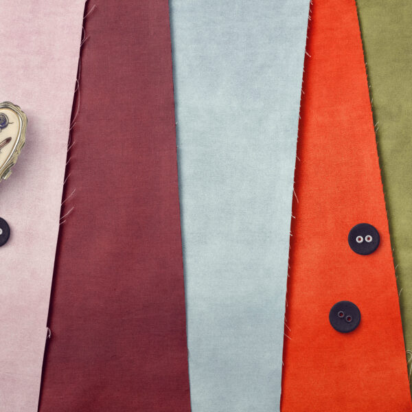 Five pieces of fabric in burgundy, light blue, orange, and olive green are arranged side by side. Two black buttons are on the orange fabric, one on the burgundy, and a tape measure is on the left.
