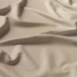 A close-up of beige fabric with a smooth texture and gentle folds, creating a wavy pattern across the surface. The material appears soft and has a subtle sheen.