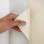 A person's hand is holding a piece of two-toned fabric, showcasing a herringbone pattern on the front. The top layer is cream-colored with the pattern, while the back appears smooth and light gray.
