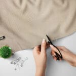 Two hands are cutting a piece of beige fabric with black scissors on a white surface. Nearby are safety pins, a clip, and a green pincushion with needles.