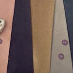 A collage of fabric swatches in muted colors including peach, dark gray, brown, and beige. Two maroon buttons are sewn onto the beige swatch, and a small, distorted clock design overlaps the peach fabric.