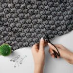 A pair of hands uses black scissors to cut black fabric with a gray elephant pattern. Nearby are safety pins and a green pin cushion on a white surface.