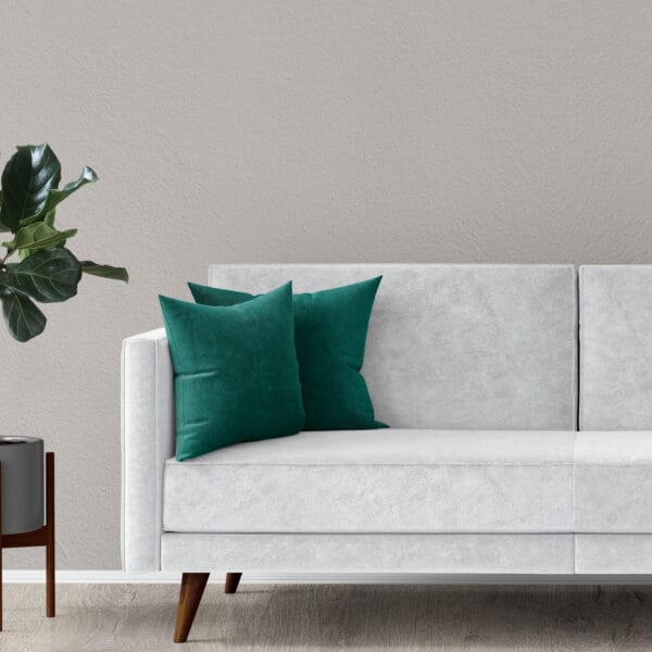 A modern white sofa with teal throw pillows sits against a light gray textured wall. A green potted plant in a gray planter on a wooden stand is positioned to the left of the sofa on a wooden floor.