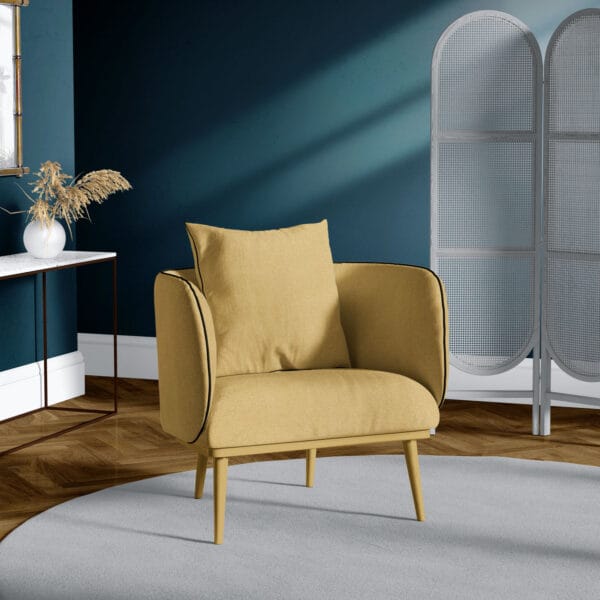 Yellow armchair with black piping on a round gray rug. Behind is a blue wall and a light gray folding screen. A white console table with dried plants and a gold-framed mirror are on the left. The floor is wooden.