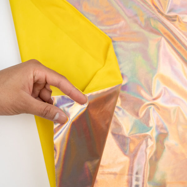 A hand lifts the corner of a shiny, iridescent sheet with a yellow underside, revealing its dual-colored layers against a plain surface.