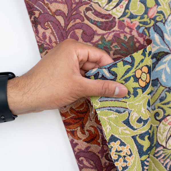 A hand holds the edge of a William Morris Pimpernel Tapestry Fabric, displaying its intricate, colorful floral and vine design, while wearing a black watch.