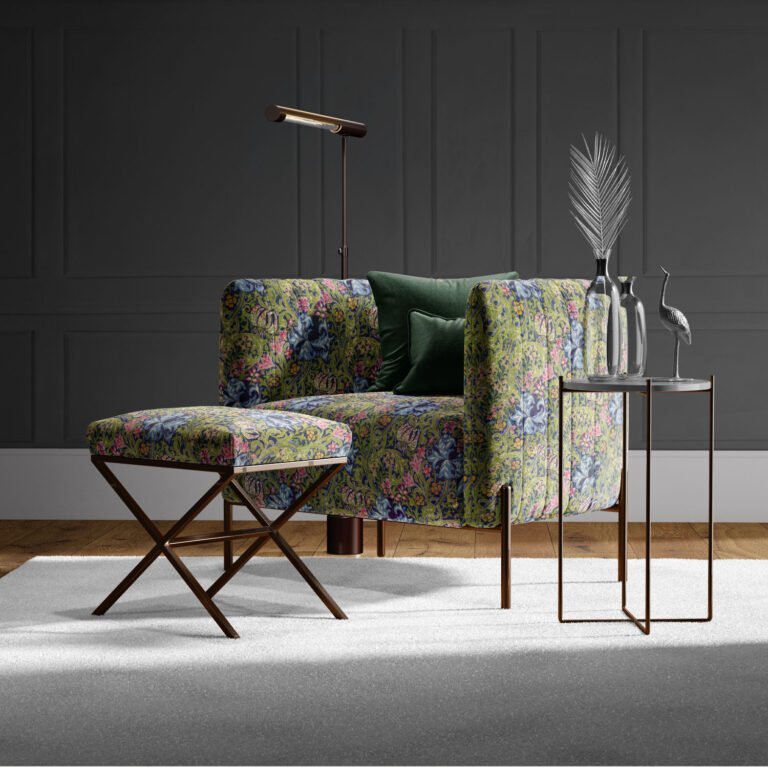A modern living room showcases a floral-patterned armchair and matching footstool featuring William Morris | Pimpernel | Tapestry Fabric, green velvet pillows, a sleek gold lamp beside a small metal side table with decorative vases, and a bird figurine against a dark paneled wall.