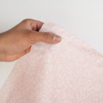 A hand holds a piece of light pink fabric with a white floral pattern against a plain white background.
