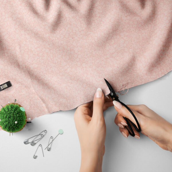A person cuts a piece of pink patterned fabric with black scissors. Nearby on a white surface are safety pins, a green pincushion with pins, and a tape measure.