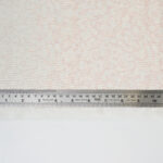 A metal ruler measuring about 16 centimeters and 6 inches lies on a sheet of pale pink floral-patterned fabric, with a strip of plain white fabric below.