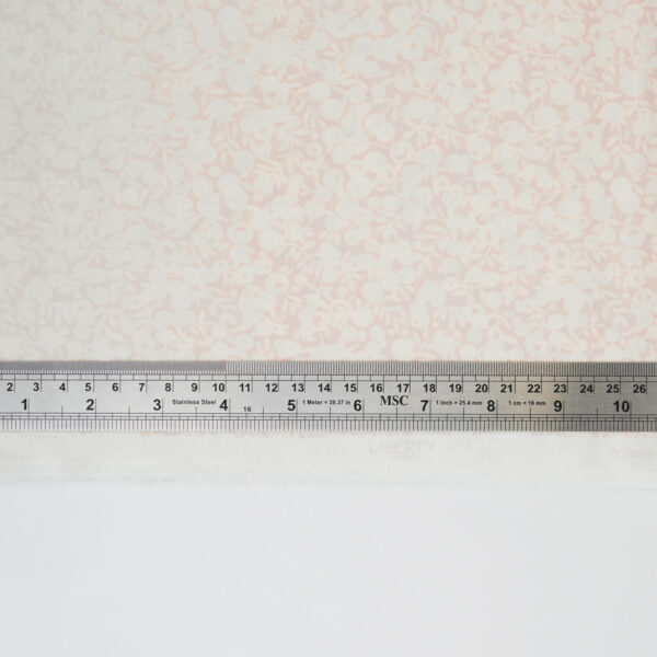 A metal ruler measuring about 16 centimeters and 6 inches lies on a sheet of pale pink floral-patterned fabric, with a strip of plain white fabric below.