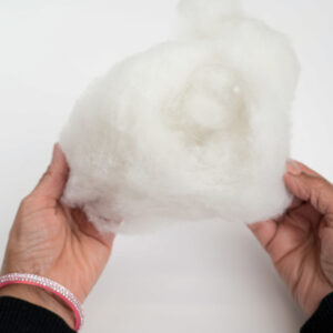Two hands holding a fluffy piece of white fiberfill or stuffing against a white background. The person on the left wears a pink and silver bracelet.
