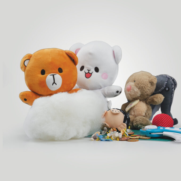 Two plush bears with exposed stuffing sit among various toys, including a brown bear, a black elephant, a red polka-dot ball, play coins, scissors, and scattered artificial flowers on a white background.