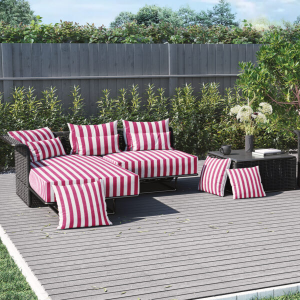 Outdoor patio set with black wicker furniture, red and white striped cushions, and pillows. A black table holds a vase, book, and cups. The area has a wooden deck, green shrubs, and a tall wooden fence.