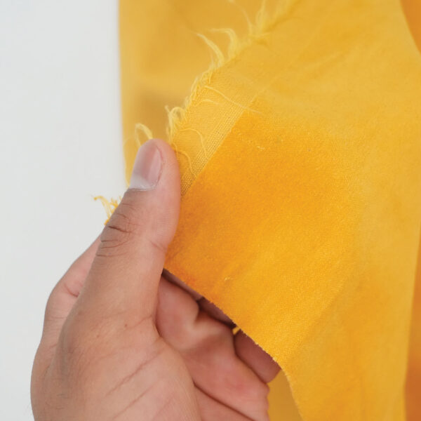A hand holding the edge of a piece of bright yellow fabric, showing a frayed, unfinished side against a plain background.