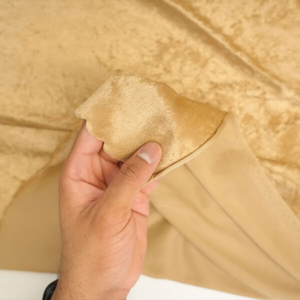A hand holds a piece of light brown velvet fabric, showing its soft texture and sheen, with more fabric spread out in the background.