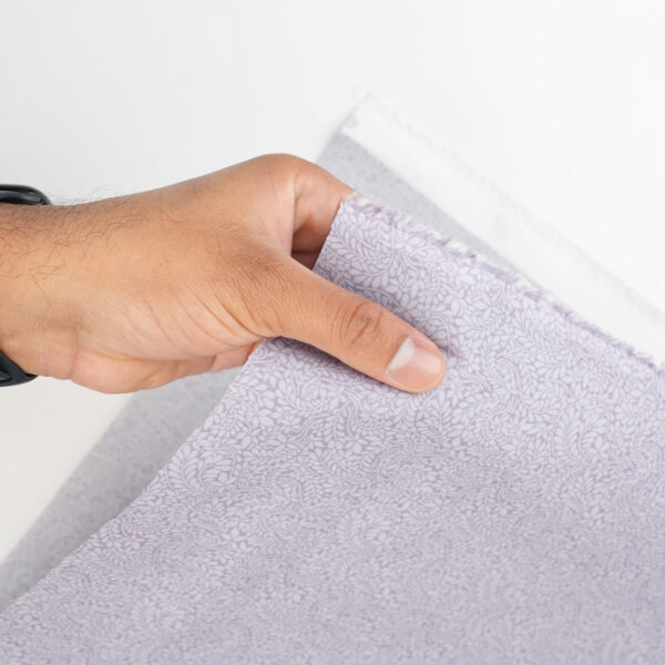A hand holding a piece of light purple fabric with a subtle, intricate leaf pattern against a white background.