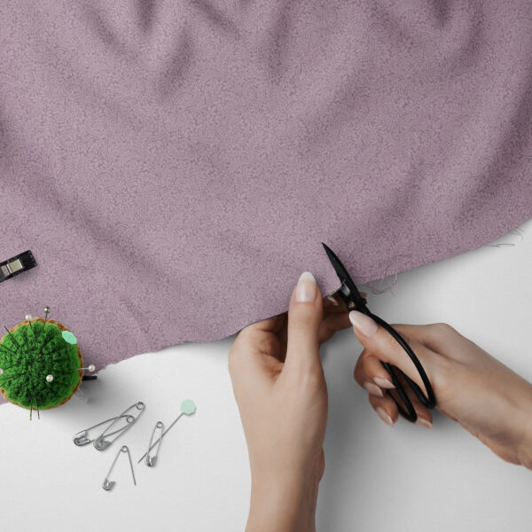 A person uses black scissors to cut the edge of a piece of light purple fabric. Nearby are safety pins, a green pincushion with needles, and sewing tools on a white surface.