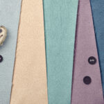 Four pieces of patterned fabric in beige, teal, blue, and purple are laid side by side. Three black buttons and a metallic thimble are placed on the fabrics. The fabric edges are slightly frayed.