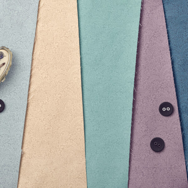 Four pieces of patterned fabric in beige, teal, blue, and purple are laid side by side. Three black buttons and a metallic thimble are placed on the fabrics. The fabric edges are slightly frayed.