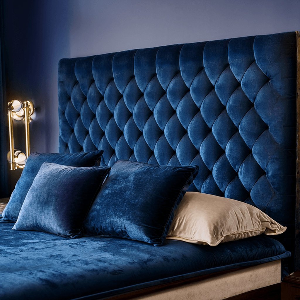 Luxurious bedroom with a large, tufted velvet headboard in deep blue, matching blue velvet bedding, and pillows, with a cream accent pillow. A modern gold lamp is visible next to the bed against a blue wall.