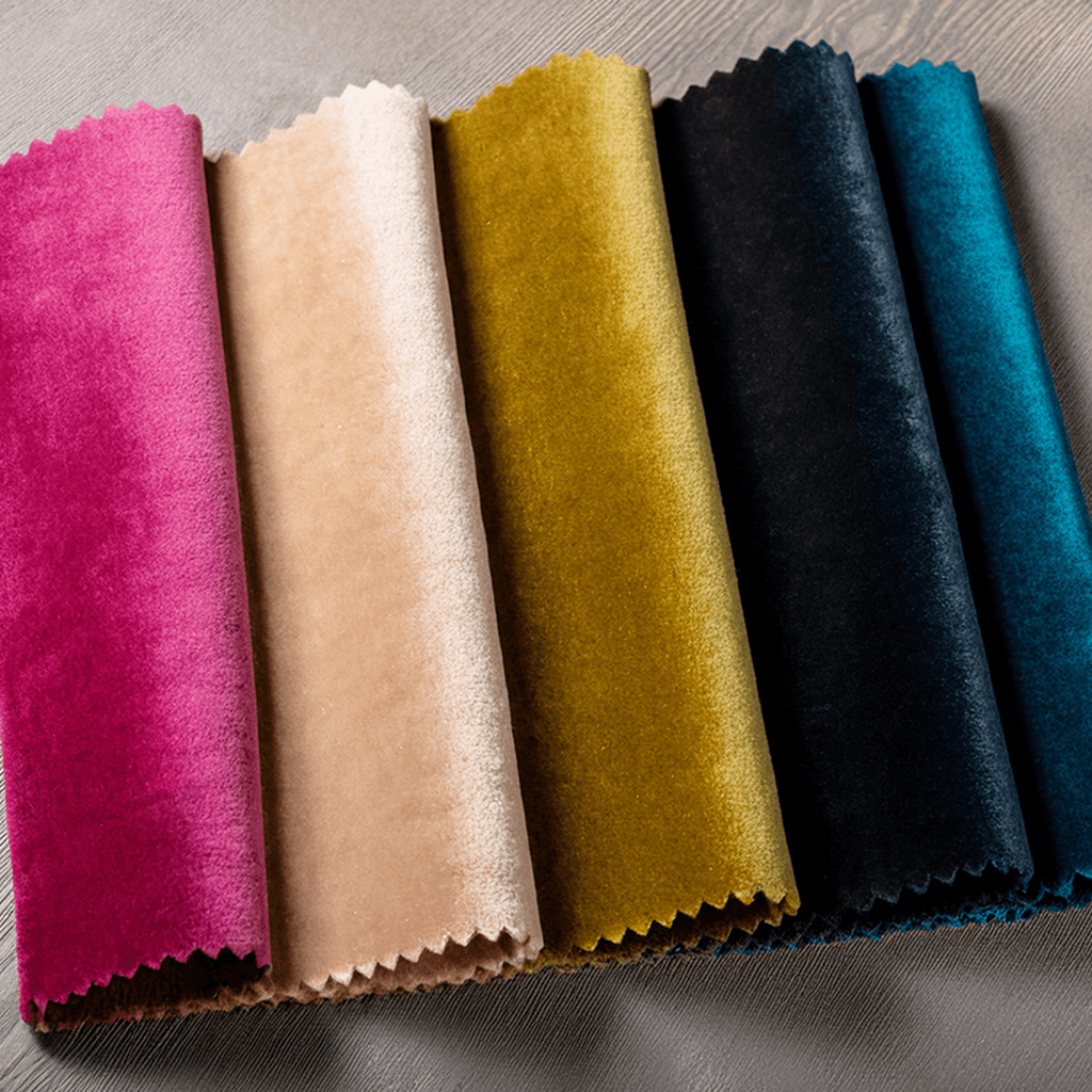 Six pieces of velvet fabric in pink, beige, gold, dark green, black, and teal are laid side by side, each with zigzag-cut edges, displayed on a light, textured surface.