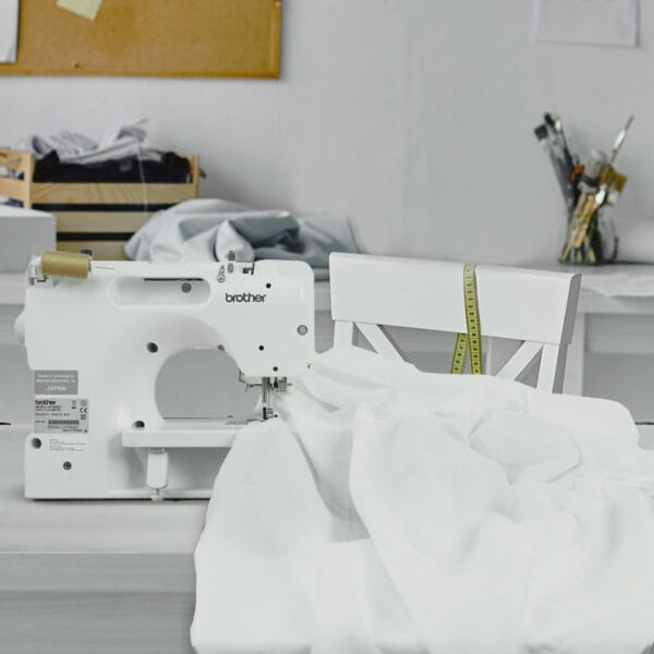 A white sewing machine on a table with white fabric being stitched, next to a white chair with a yellow measuring tape draped over it; the workspace includes art supplies and fabric in the background.