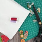 A piece of white fabric, red and purple thread spools, scissors, floral pins, decorative buttons, and a green cutting mat on a wooden surface, with artificial flowers scattered nearby.