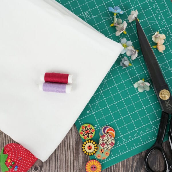 A piece of white fabric, red and purple thread spools, scissors, floral pins, decorative buttons, and a green cutting mat on a wooden surface, with artificial flowers scattered nearby.